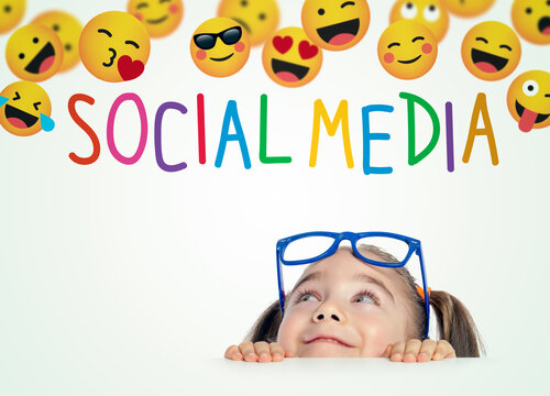 Beautiful cute little girl with eyeglasses hiding behind a  table and looking at group of emojis and Social Media text above her head.