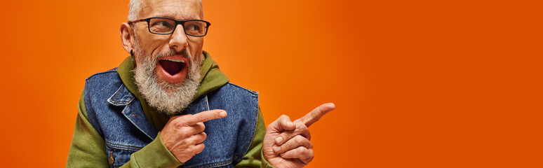 joyful handsome mature man in trendy denim vest posing on orange backdrop and pointing away, banner