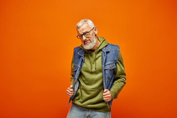 attractive fashionable mature man in vibrant outfit posing on orange backdrop and looking at camera