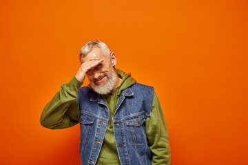joyful good looking mature man in vivid attire with gray beard smiling at camera on orange backdrop