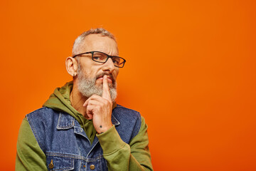 handsome focused mature man in green hoodie with glasses and gray beard posing on orange background