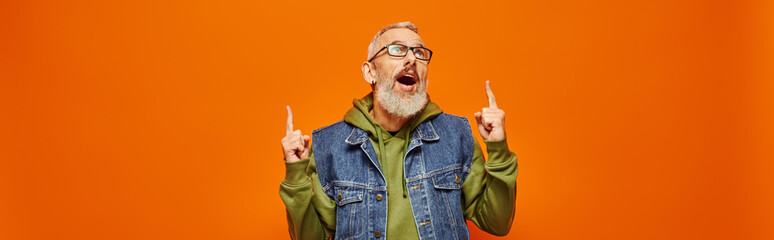 surprised mature man with glasses and beard in vivid hoodie pointing up on orange backdrop, banner