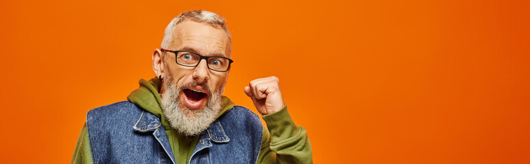 jolly mature man in green vivid hoodie posing with fists near face on orange background, banner