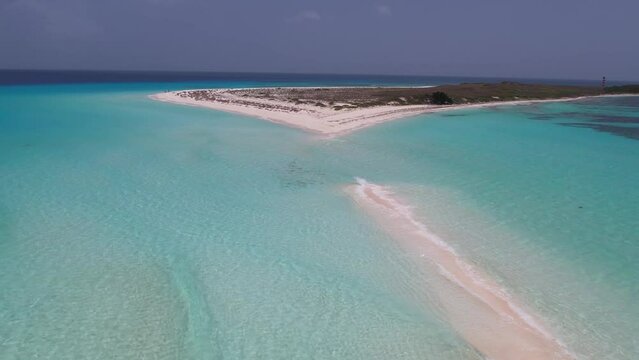 Aerial pullback above sand island with crystal clear light blue ocean water waves