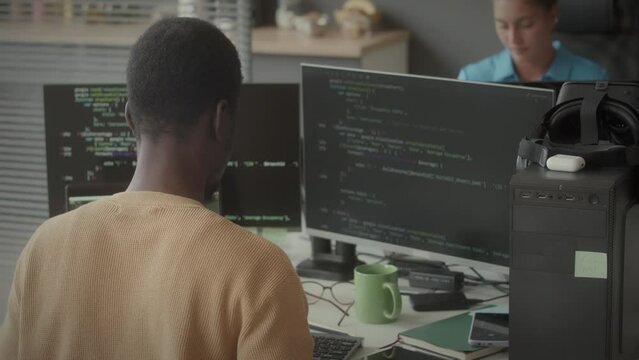 View from behind of Black male programmer sitting at office desk and working on computer with dual monitor while creating source code