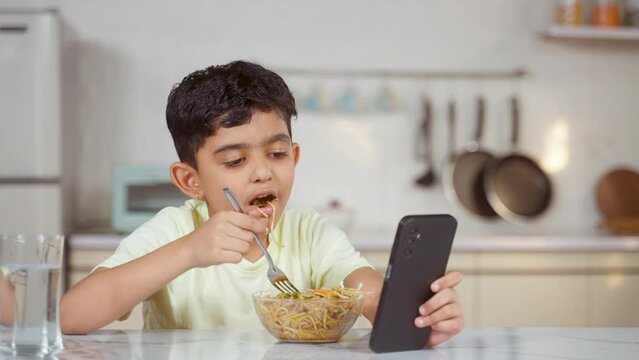 Indian preteen kid eating noodles while using mobile phone at home on dining table - concept of unhealthy lifestyle, internet addiction and technology