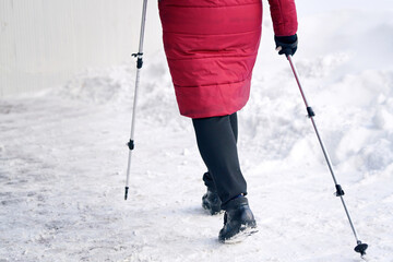 Senior woman walking along slippery snowy sidewalk with walking poles. Old woman practicing Nordic...