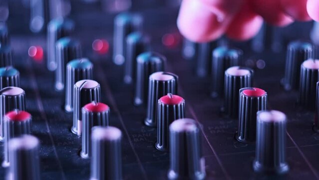 Male hand adjusting buttons on audio mixer in neon light close-up. Working mixing console in colorful background. DJ plays music at night club in multi-color effect. Sound engineer moving faders level