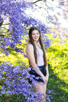 Happy long haired teen girl standing amongst purple jacaranda flowers in park outside