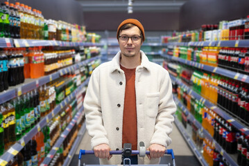 Male shopper with glasses and beanie holding a cart in a drink aisle. Portrait of a man with a...