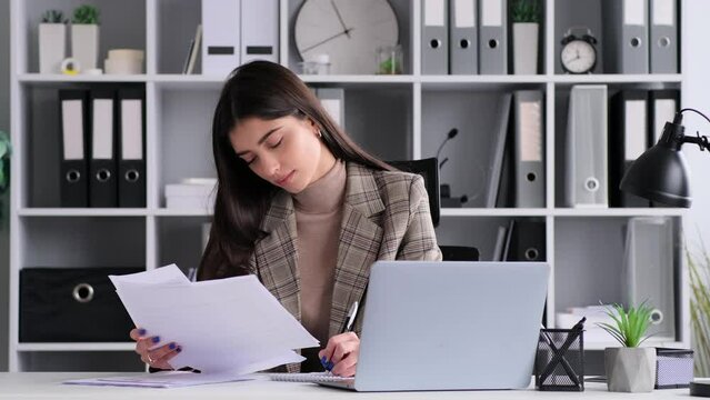 Positive Caucasian woman working in the office with papers, reports, fees sitting at the table in the office. Analyzing documents, accounting business.