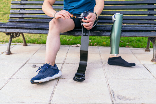 Focused male athlete sitting on a bench in a park, adjusting a high-tech carbon fiber sports prosthetic leg, preparing for an outdoor workout.