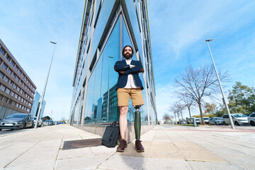 Self-assured businessman with artificial leg confidently standing outside a reflective glass building, arms crossed.