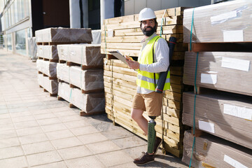 Construction expert in safety vest assesses materials for quality control beside stacked pallets.