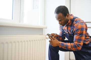 Man in workwear overalls using tools while installing or repairing heating radiator in room