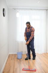 Professional cleaner in blue uniform washing floor and wiping dust from the furniture in the living room of the apartment. Cleaning service concept