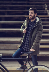 Bearded men smiling wearing a green jacket and shirt standing on the stairs holding the wheel of a vintage bicycle and looking arround. Stairs behind him in the background. Outdoor shot.