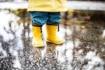 Small infant boy wearing yellow rubber boots and yellow waterproof raincoat standing in puddle on a...