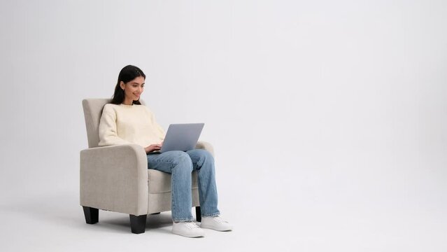 Positive Caucasian woman browsing on a laptop while sitting in a armchair on a white background. Online connection, internet communication, modern technology, freelance work concept.