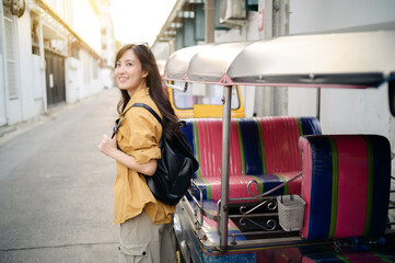 Young Asian woman backpack traveler standing a side of Tuk Tuk taxi on summer vacations at Bangkok, Thailand. Journey trip lifestyle, Asia summer tourism concept.