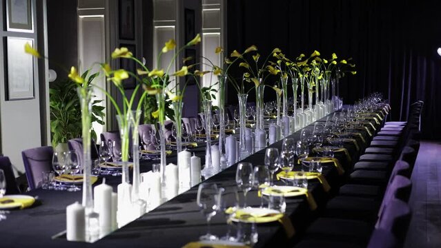 Festive dinner table served with dishes and cutlery at both sides and decorated with yellow callas flowers and candles in middle. Dark interior restaurant in violet and black colours hosting party.