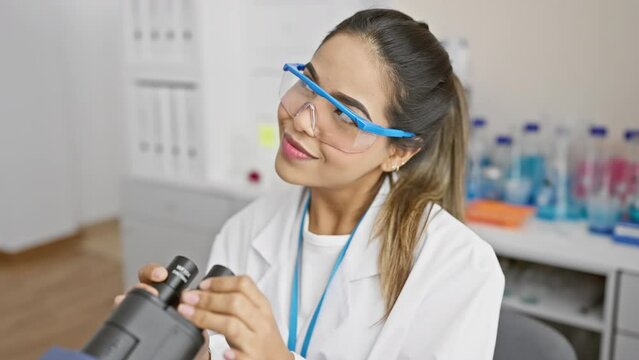 Cheerful young latin woman scientist thoughtfully looking away into space, smiling as she works with a microscope indoors at the lab, hopeful for a positive result, her curiosity piqued.