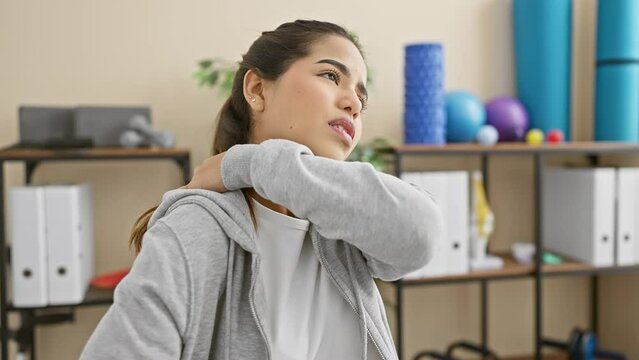 A young hispanic woman in a rehabilitation clinic expresses discomfort while holding her neck, indicative of a possible injury or pain.