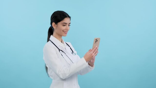 Positive female doctor uses a phone on a blue background. Technology and healthcare blend, online medical service, modern medicine, medical staff concept.