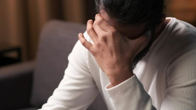 Sad Indian man suffers from headache, migraine, in the evening on the sofa in the living room at home. Bad health, restless businessman, illness, tiredness.