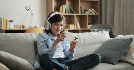 Teenage caucasian boy wearing headphones playing video game on phone sitting on sofa at home. Domestic leisure activity time, mobile video game, online entertainment.