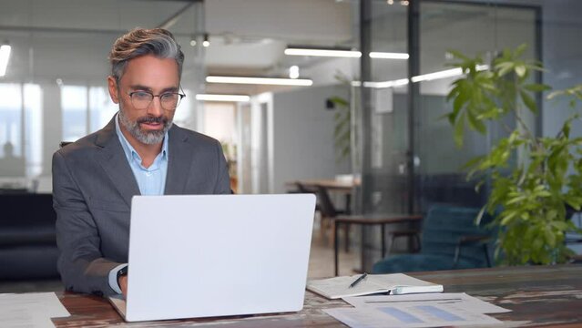Serious mature European business man ceo trader using computer, typing, working in modern office, doing online data market analysis, thinking planning tech strategy looking at laptop with copy space.