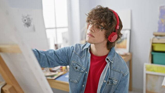 A thoughtful young man with glasses and headphones engaged in painting in a bright art studio.