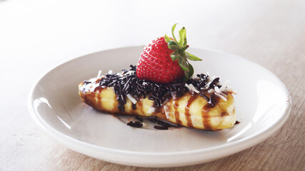 grilled banana adorned with a delectable ensemble of chocolate, grated cheese, and fresh strawberries, elegantly presented on a plate a top a rustic wooden table.