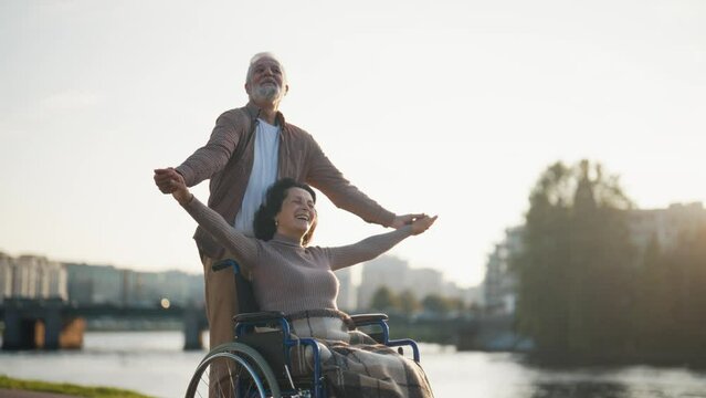 Elderly husband walking with wife in wheelchair. Senior family couple spending time in city park. Man caring woman showing love, they look happiness. Living with physical disability, freedom concept.
