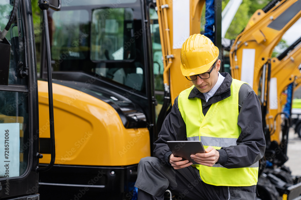 Poster Engineer in a helmet with a digital tablet stands next to construction excavators..