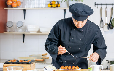Portrait close up handsome professional Japanese male chef wearing black uniform, hat, cooking, making takoyaki street food, smiling with confidence, standing in kitchen. Restaurant, Hotel Concept.