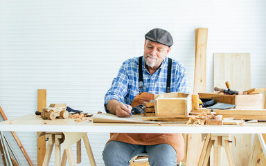 Senior Caucasian happy retired old male woodworker or carpenter smiling, wearing check shirt with apron, vintage hat, creating DIY wooden mini figure toy for decoration as hobby after retirement.