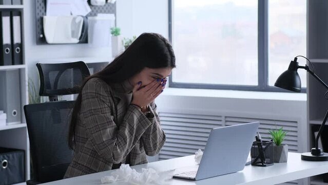 Illness Caucasian woman sneezing and blowing runny nose at napkin while working with laptop in office. Infection or allergy, grippe, respiratory disease.