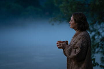 A woman in a sweater on the bank of a foggy river in the evening..