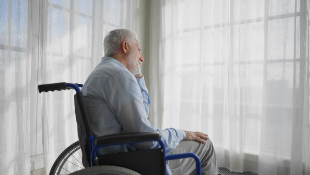 Old man in wheelchair looks at window. Thoughtful gray haired senior male at home enjoying pastime leisure. Living with physical legs trauma paralysis, melancholy, special needs people concept.