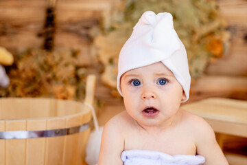 a cute little child in a towel and a turban on his head after bathing sits in a steam room in a...