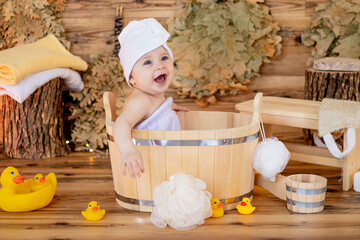 a cute little child in a towel and a turban on his head after bathing sits in a basin in a steam...