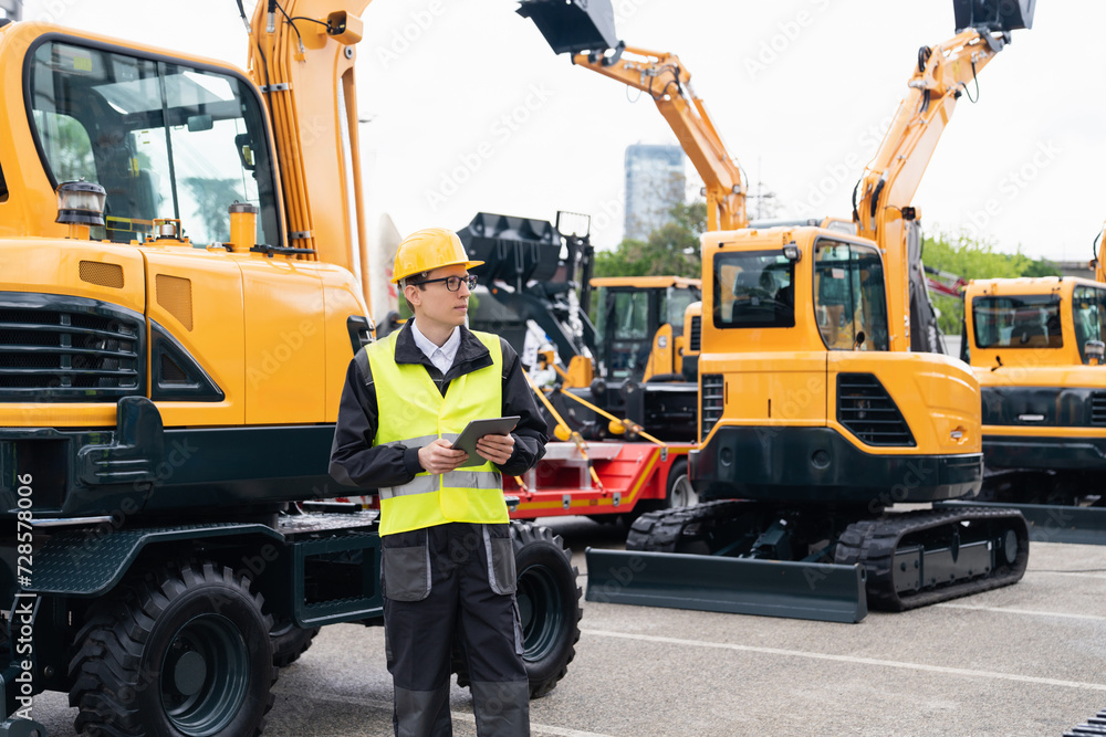 Poster Engineer in a helmet with a digital tablet stands next to construction excavators..