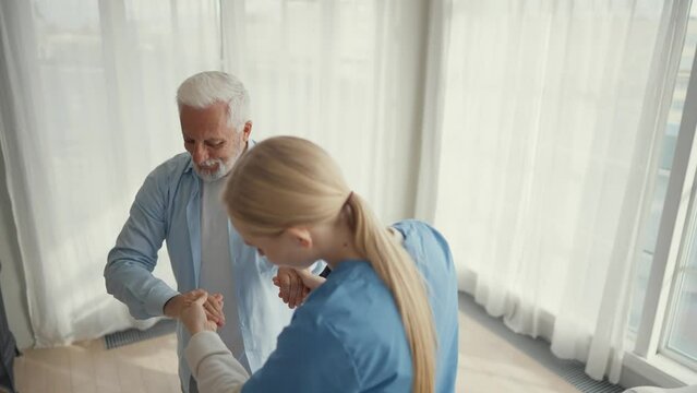 Nurse helps old wheelchair man to stand up. Physical therapist woman provides medical help support to senior paralyzed male giving hands. Palliative care in late stage neurodegenerative pathologies.