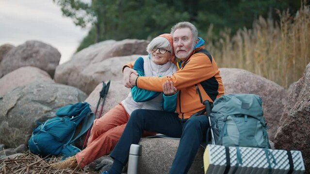 Senior happy family couple hugging talking together admiring nature sitting having break on stones. Hikers old man woman resting cuddling enjoying activity on nature. Hiking and travel concept.
