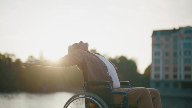 Senior man in wheelchair enjoys freedom. Paralysis male with special need spreading arms on sides smiling breathing fresh air walking in city park. Living with physical disability in old age concept.