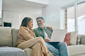 Happy middle aged couple using laptop computer relaxing on couch at home. Smiling mature man and woman talking having fun laughing with device sitting on sofa in sunny living room. Candid shot.