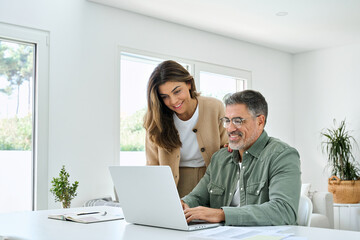 Smiling middle aged senior man working on computer sitting at table with wife standing nearby in...