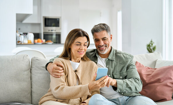 Happy older mature senior family couple, middle aged man and woman looking at cell phone using smartphone mobile technology device together sitting at home relaxing on couch doing online shopping.