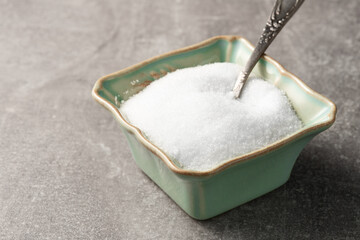 White granulated sugar in a turquoise sugar bowl with a metal spoon on a gray textured background.